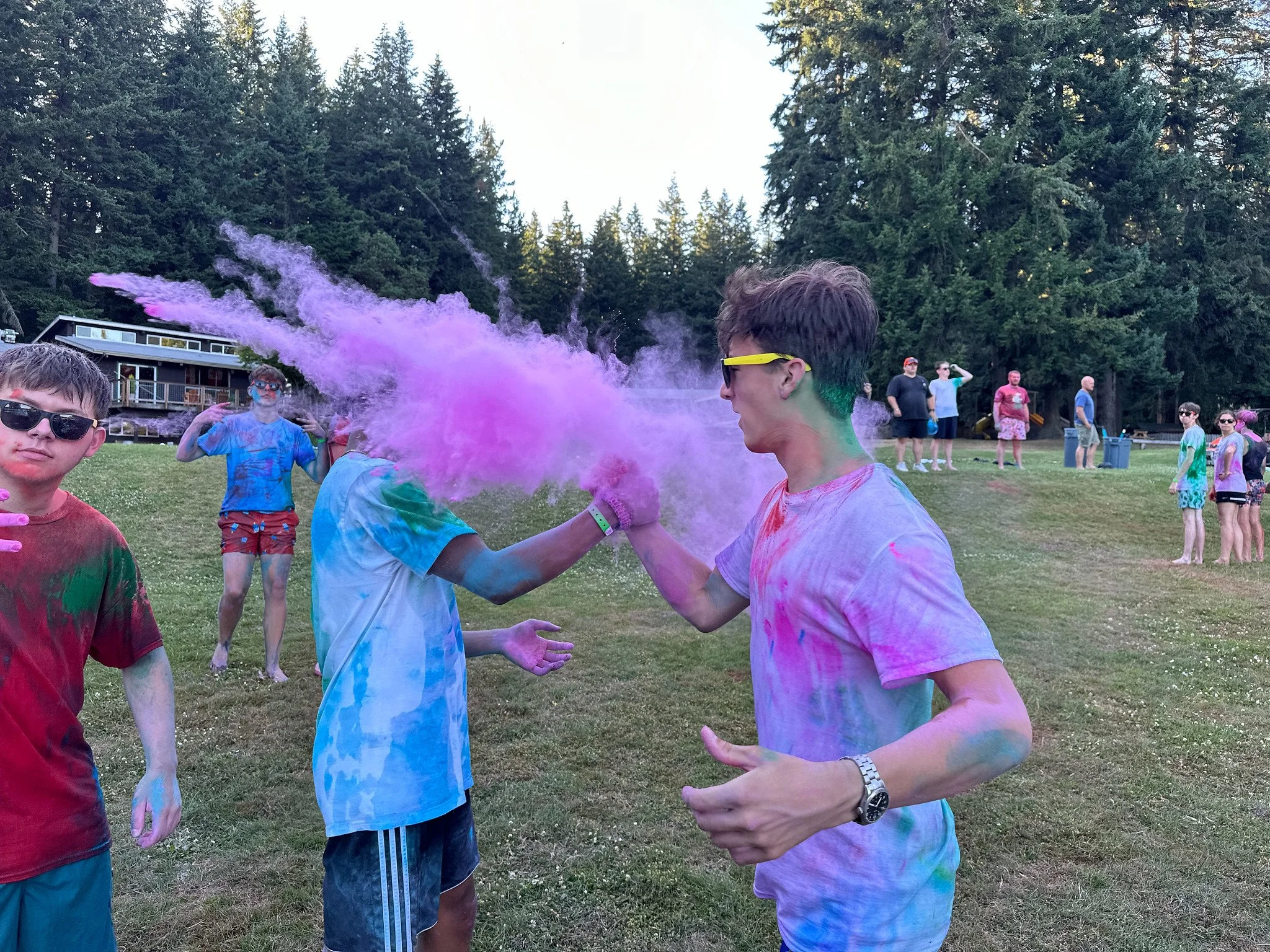 People participating in a color powder event outdoors, with one person throwing purple powder, while others are covered in various colored powders, trees in the background, and a small building.