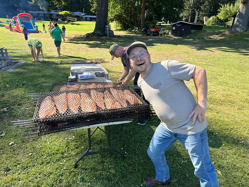 People at an outdoor barbecue event with salmon on the grill, a man posing in front, and a playground with an inflatable slide in the background.