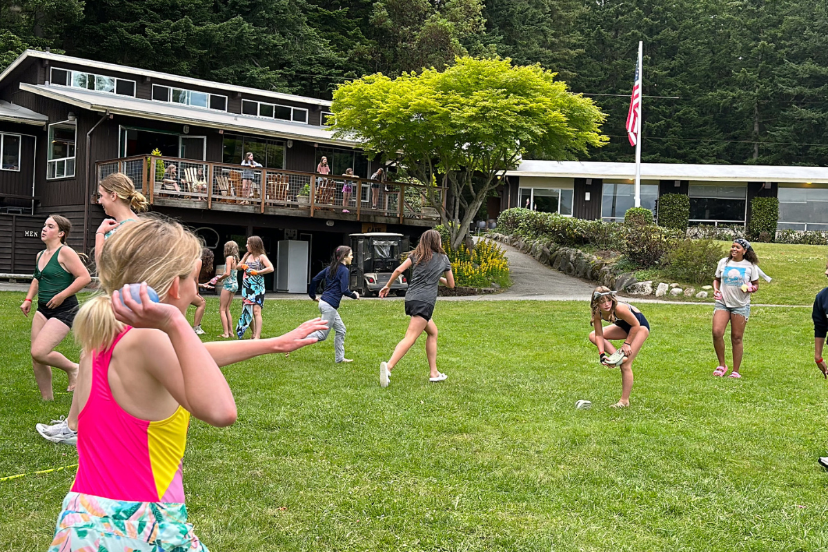 Group of children playing a game on a grassy field, wooden building in the background, surrounded by trees, American flag visible.