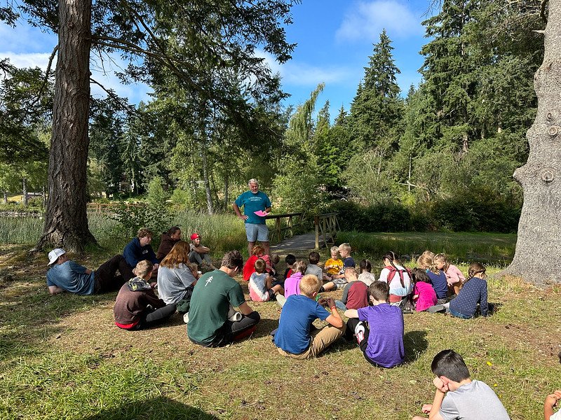 A group of children and a few adults sit on grass in a forested area, listening to a person standing and speaking in front of them. The scene is outdoors with trees, grassy ground, and a bridge visible in the background.