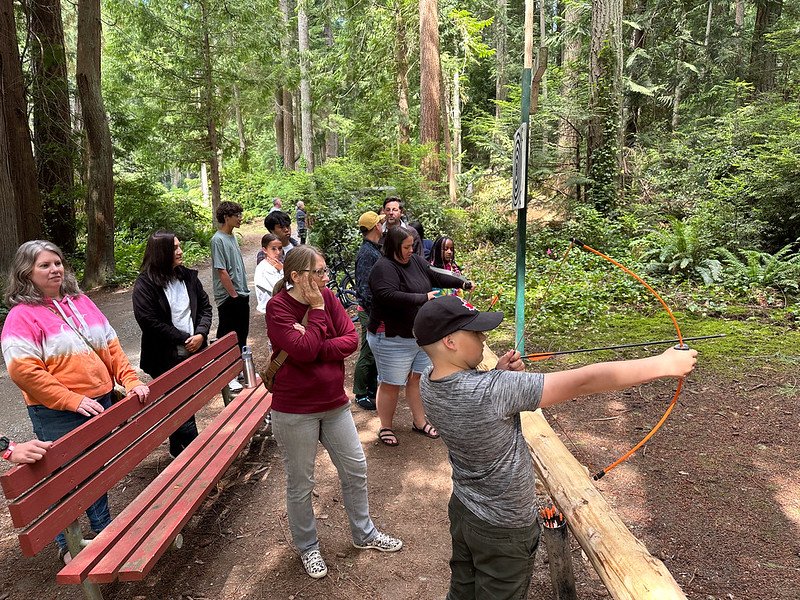 A group of people watching a child practicing archery in a forest setting.