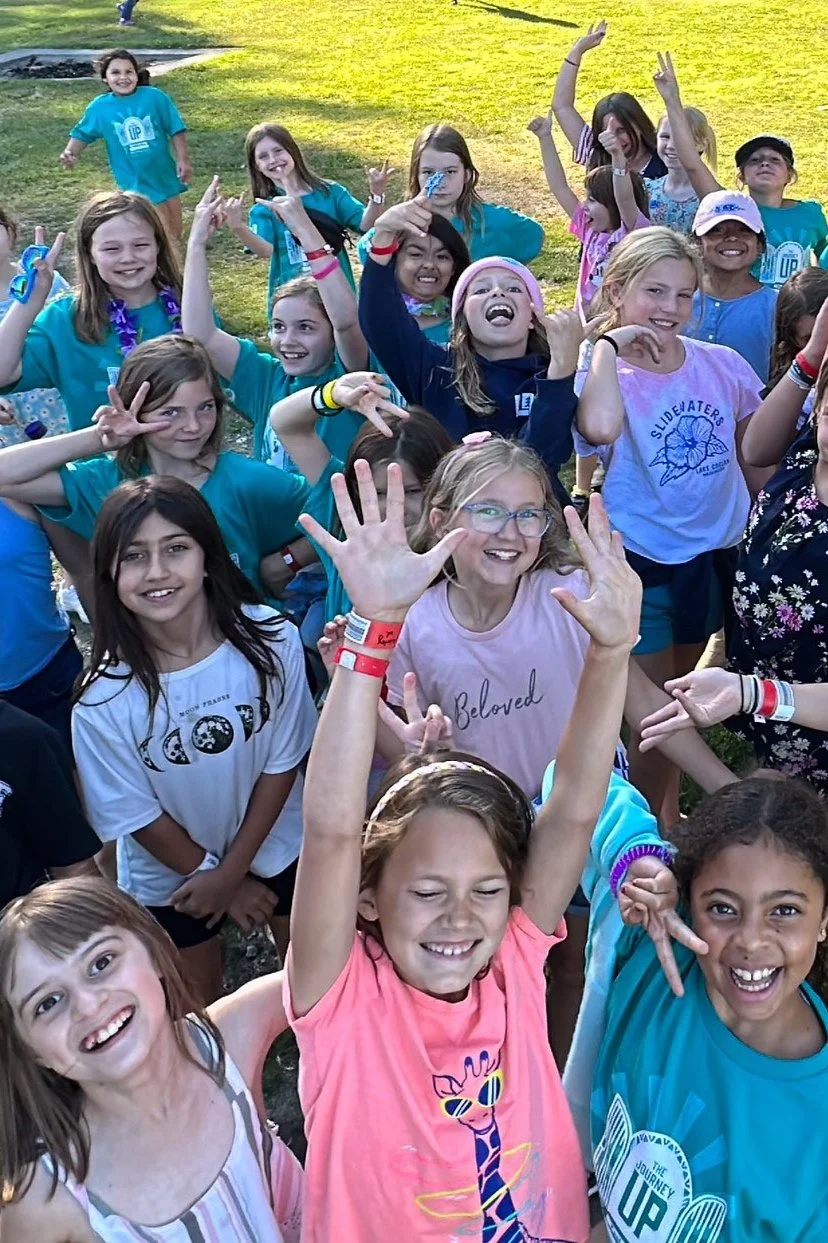 A group of smiling children enjoying an outdoor event, some making peace signs and playful gestures, on a grassy field during daytime.