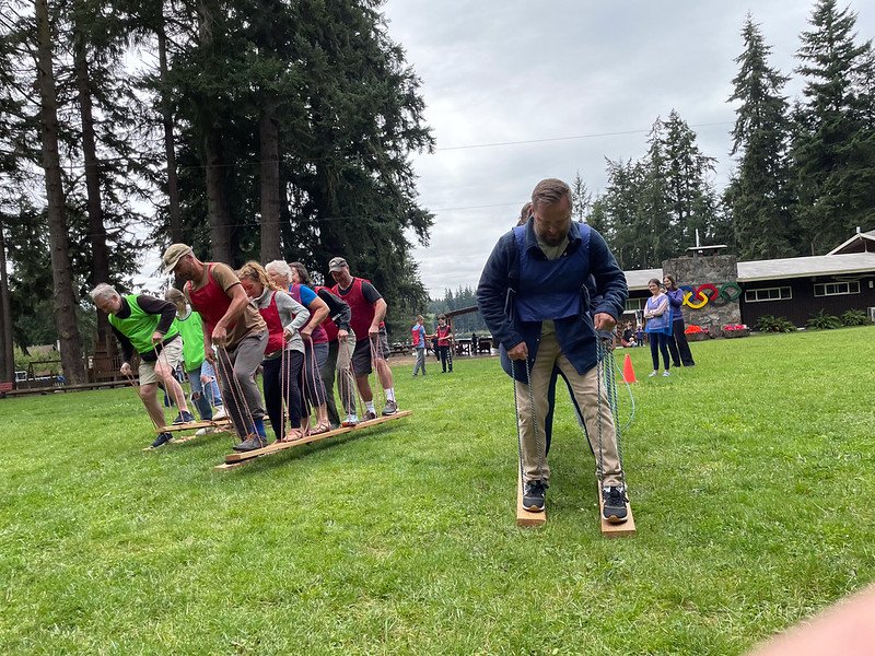 Group of people participating in an outdoor team-building activity using wooden planks on a grassy field, surrounded by trees and buildings in the background.