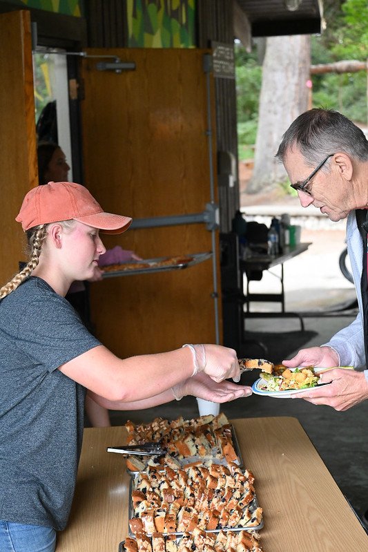 Person serving food at an outdoor event, with multiple loaves of bread on a table and a person receiving a plate of salad.