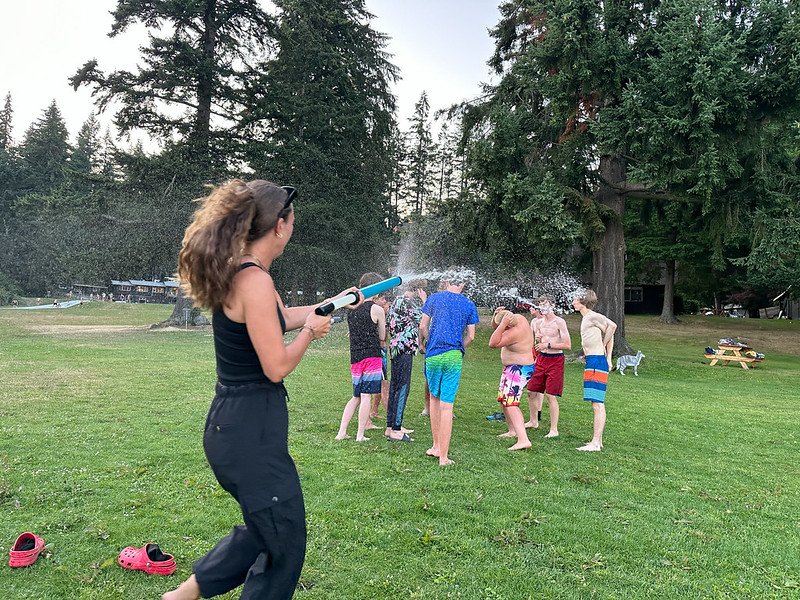 Group of people playing with water outside on a grass field, using a water gun, surrounded by trees.