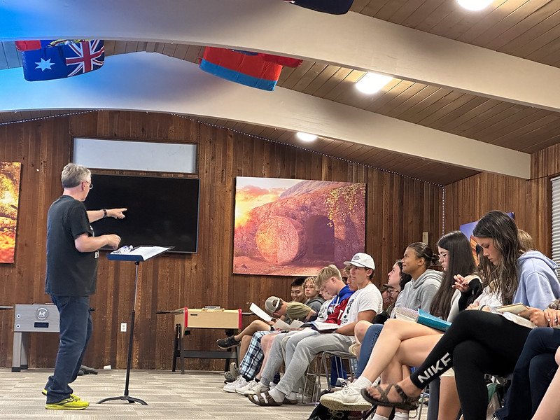 Man speaking to a seated group in a wooden room with flags on the ceiling and a large screen on the wall.