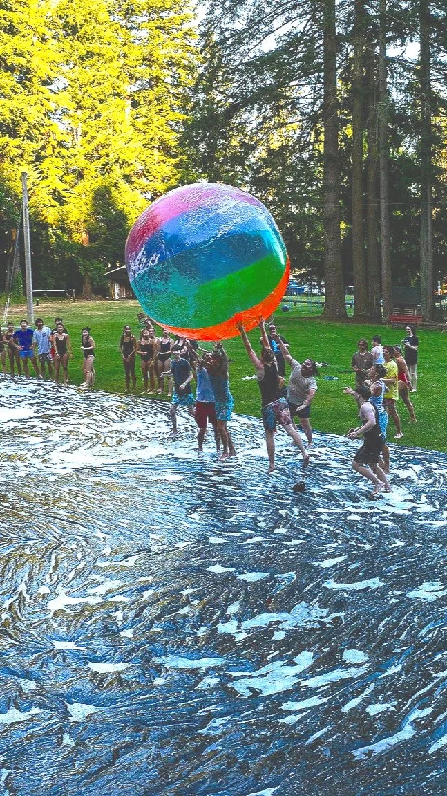 People at a splash pad playing with a large, colorful beach ball