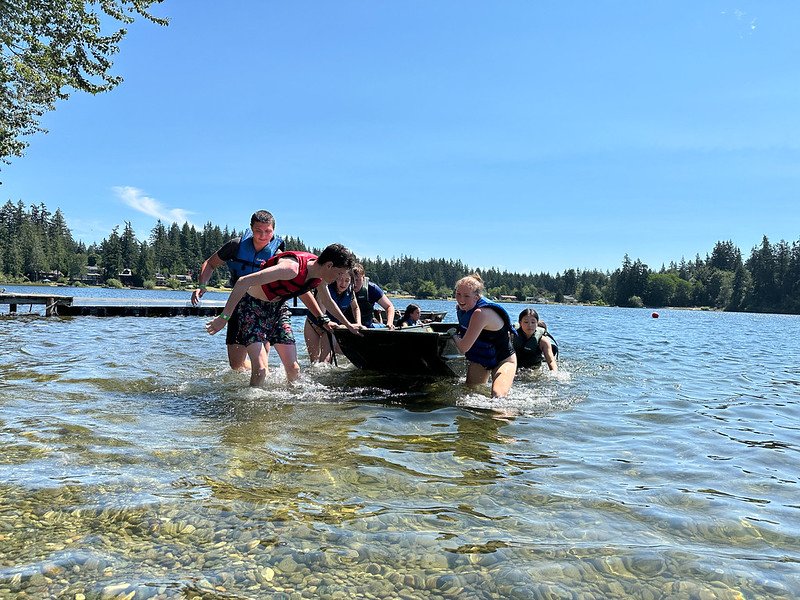 Group of people in life jackets wading in a lake, pushing a small boat, with a forested shoreline in the background.