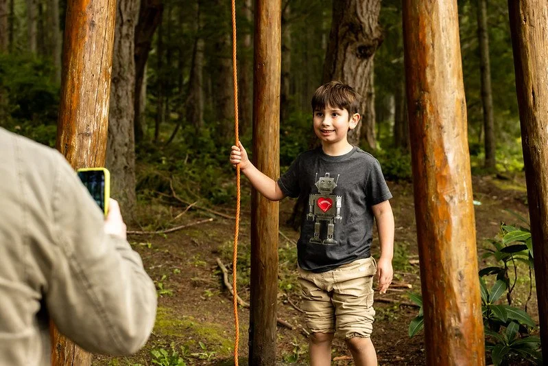 Child standing among wooden posts holding a rope, wearing a robot-themed t-shirt and shorts, while someone photographs with a smartphone in a forest.