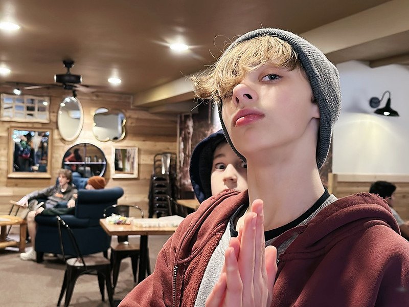 Teenager wearing a beanie and red hoodie posing with hands together in a cozy room with wooden walls.