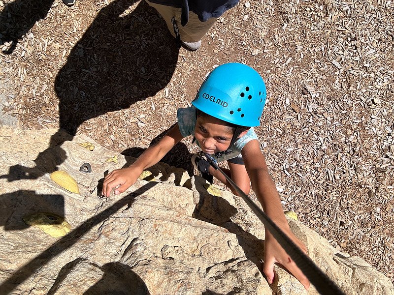Child rock climbing outdoors wearing a blue helmet with a safety rope.