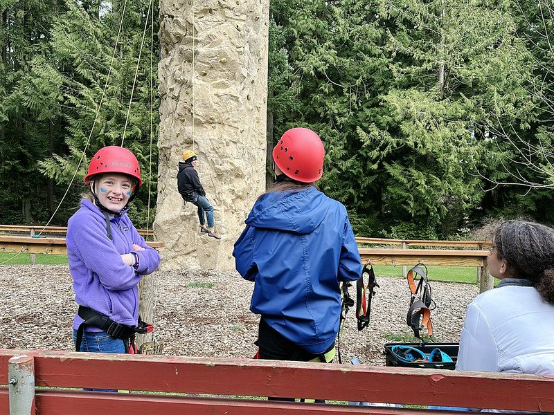 Group of children wearing helmets and safety gear near a climbing wall in an outdoor park setting.