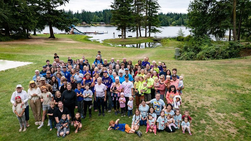 Large group gathered outdoors near lake, surrounded by trees, possibly for family or community event; diverse ages.