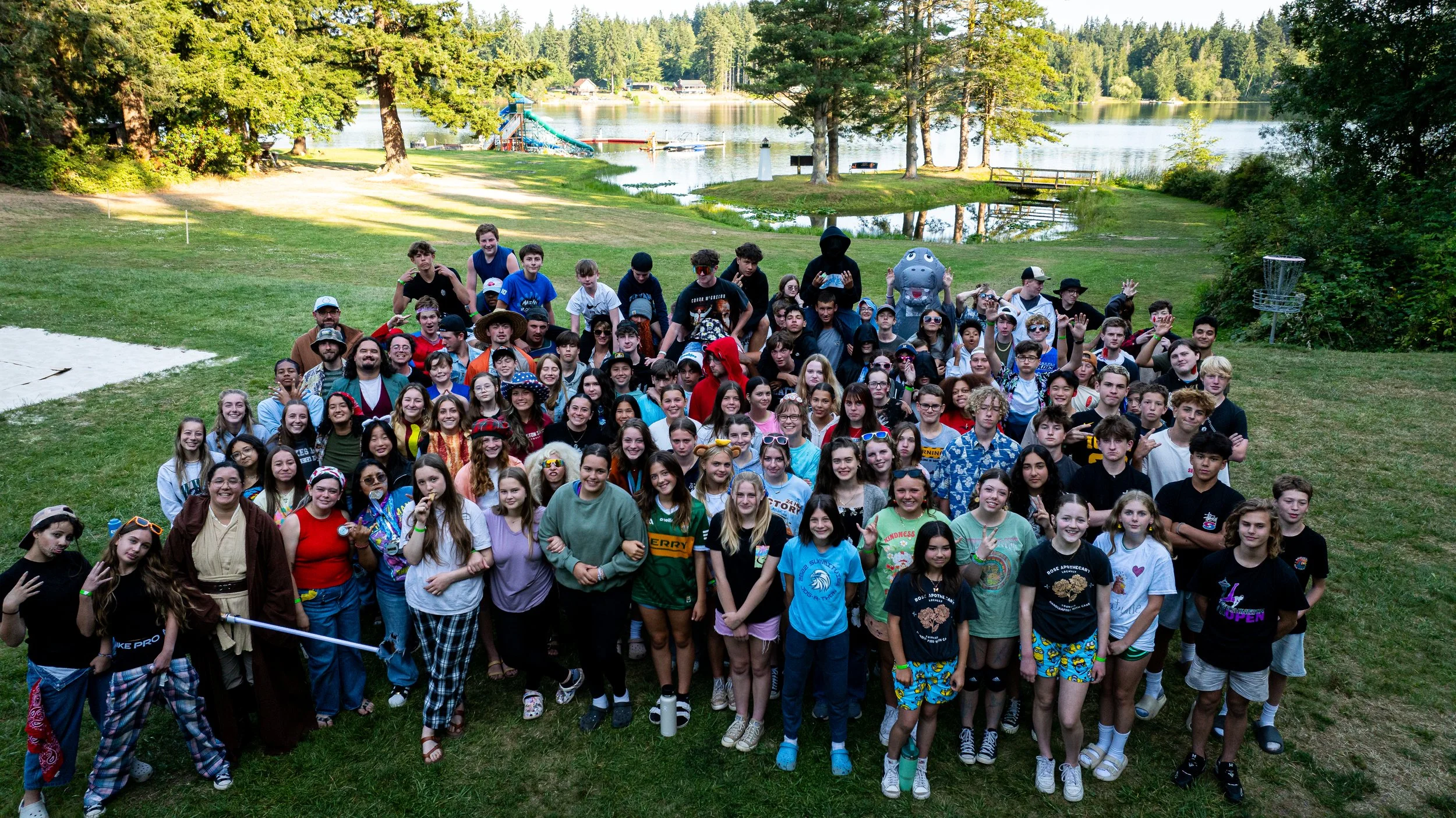 A large group of children and teenagers gathered outdoors on a grassy area near a lake, with trees and a playground in the background. Some are dressed in costumes and casual clothing, smiling and posing for a group photo.