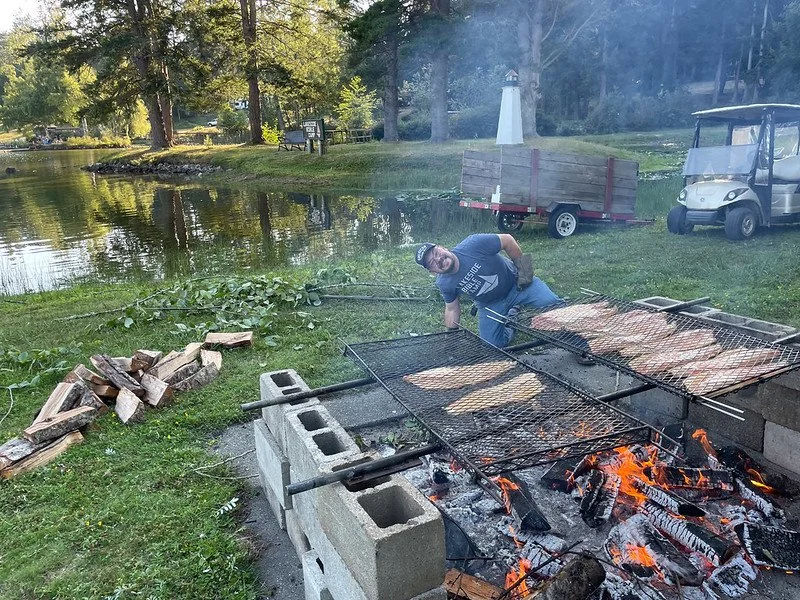 Outdoor barbecue scene with man grilling meat over fire pit, surrounded by nature with trees and a pond in the background.