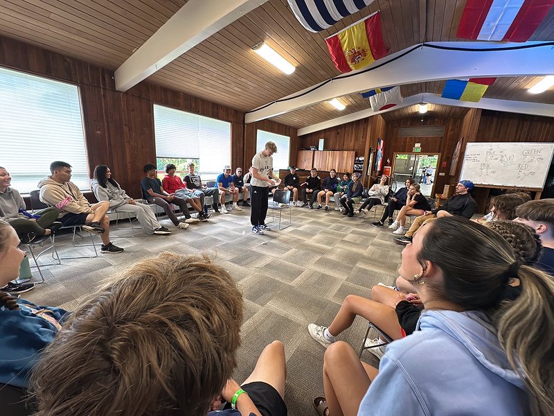 A group of people sitting in a circle in a room with wood-paneled walls, large windows, and international flags hanging from the ceiling. One person is standing in the center addressing the group. The room has a casual, meeting-like atmosphere.