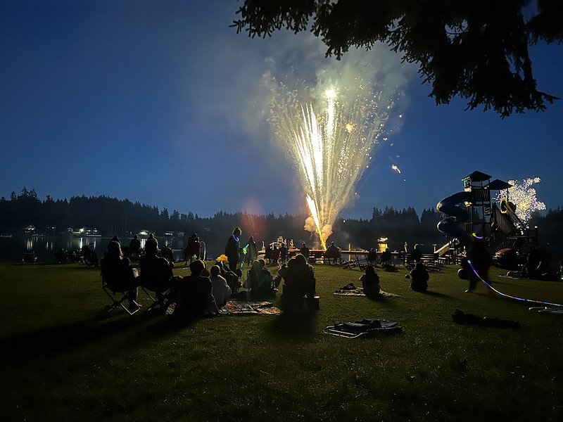 Evening outdoor gathering with fireworks display over a lake, people sitting on blankets on grassy area, playground in the background, and trees along the horizon.