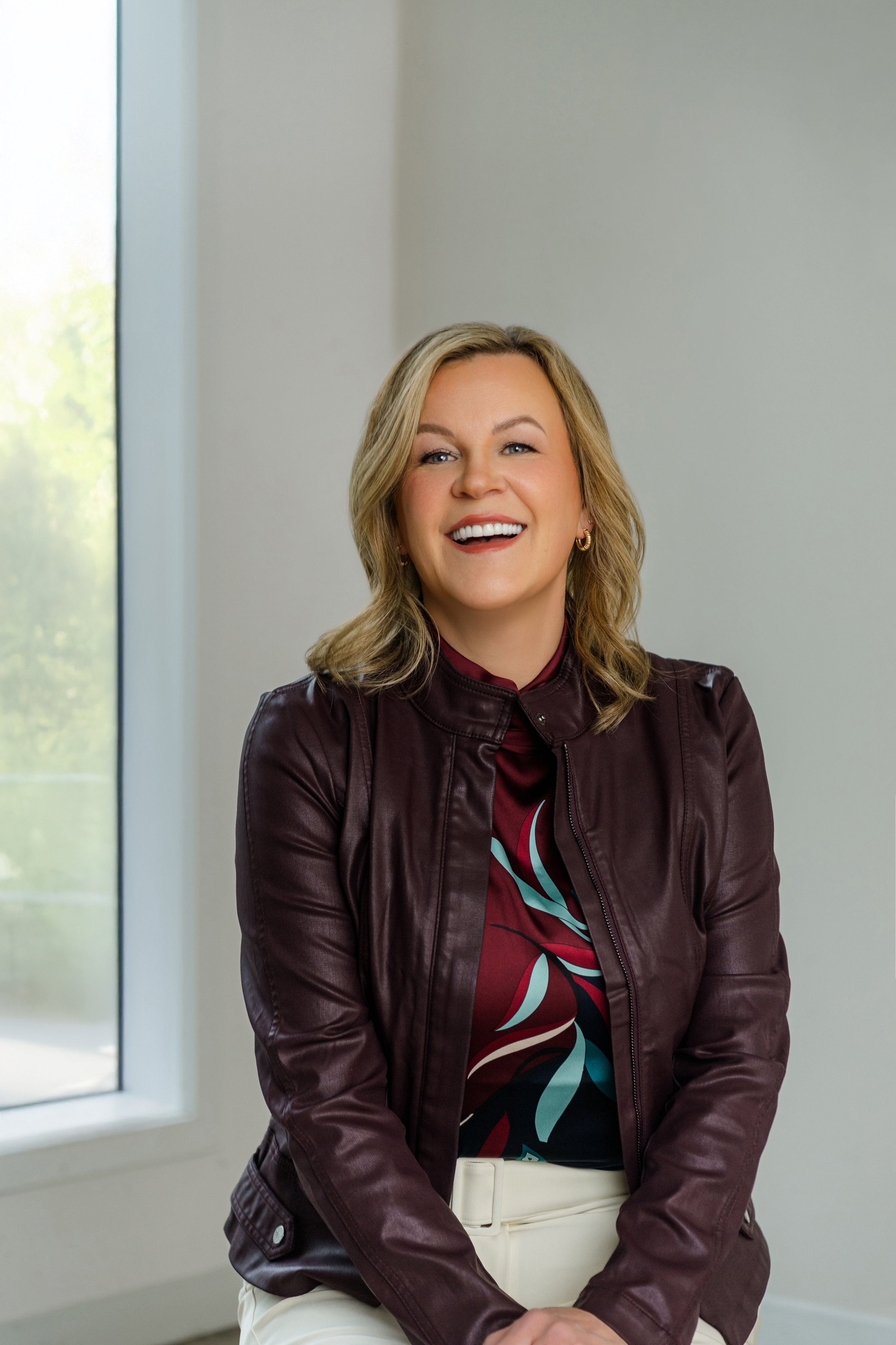 Julie smiling and sitting indoors near a window, wearing a dark leather jacket, patterned blouse, and white pants.