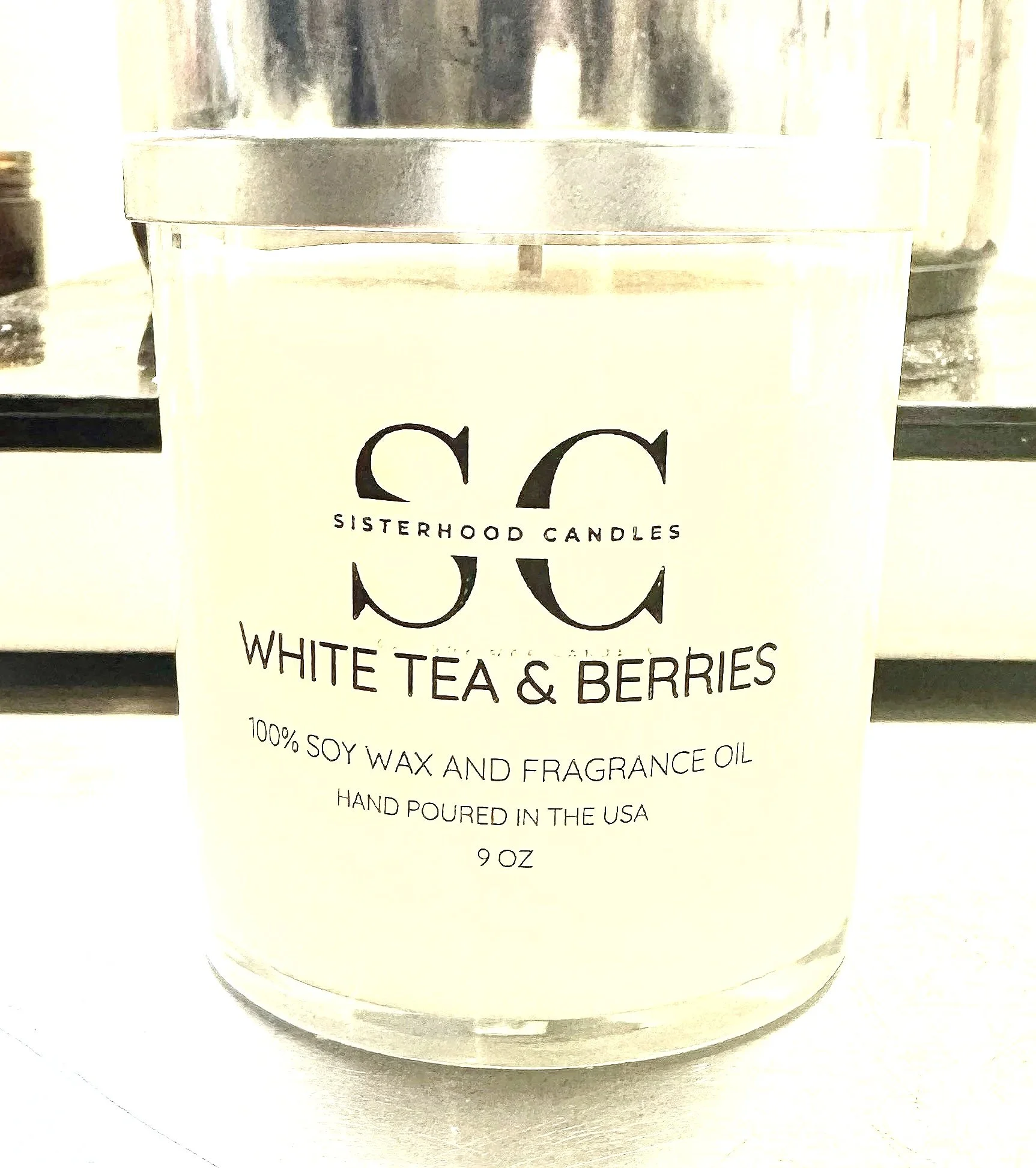 A white candle in a glass jar with a silver lid, labeled "Sisterhood Candles White Tea & Berries, 100% soy wax and fragrance oil, hand poured in the USA, 9 oz."
