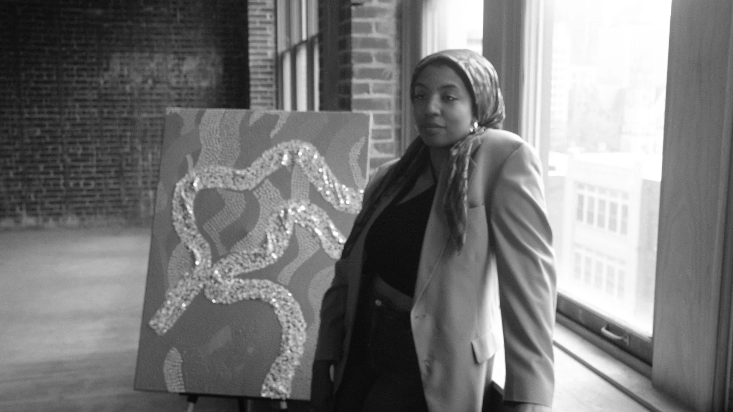 Trayvia Iman in a headscarf and blazer stands next to an abstract painting on an easel in a loft with brick walls and large windows. Downtown St. Louis. The painting features swirling patterns.