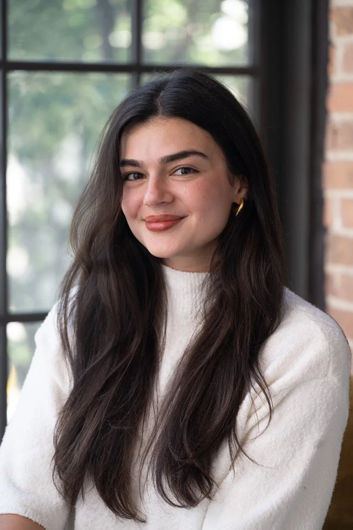 A young woman with long, wavy dark hair, wearing a white sweater and gold hoop earrings, smiling in front of a window with a blurred outdoor background.
