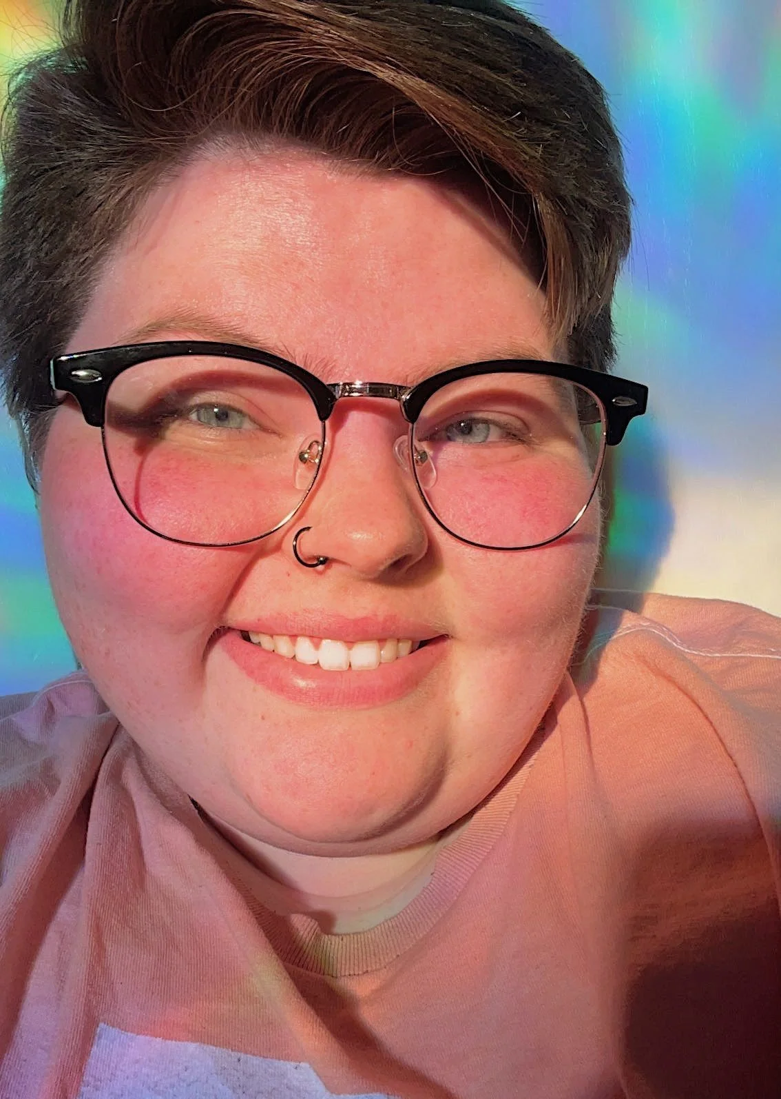 Close-up photo of a person with short brown hair, wearing round glasses with black frames, smiling, with light skin and a septum piercing, in front of a colorful rainbow-like background.