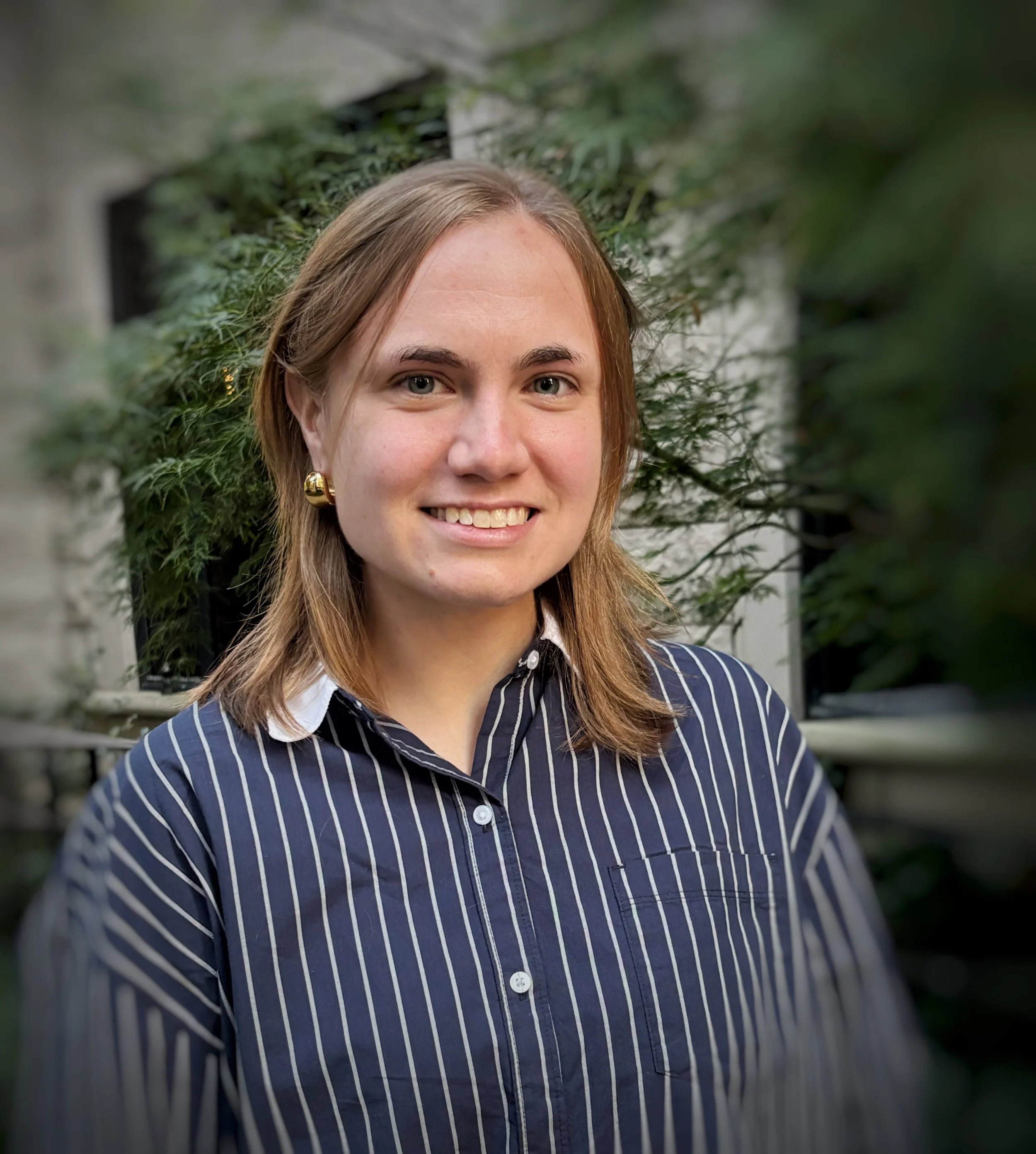A woman with shoulder-length light brown hair, wearing a navy blue and white striped button-up shirt and gold earrings, standing outdoors with green foliage in the background.