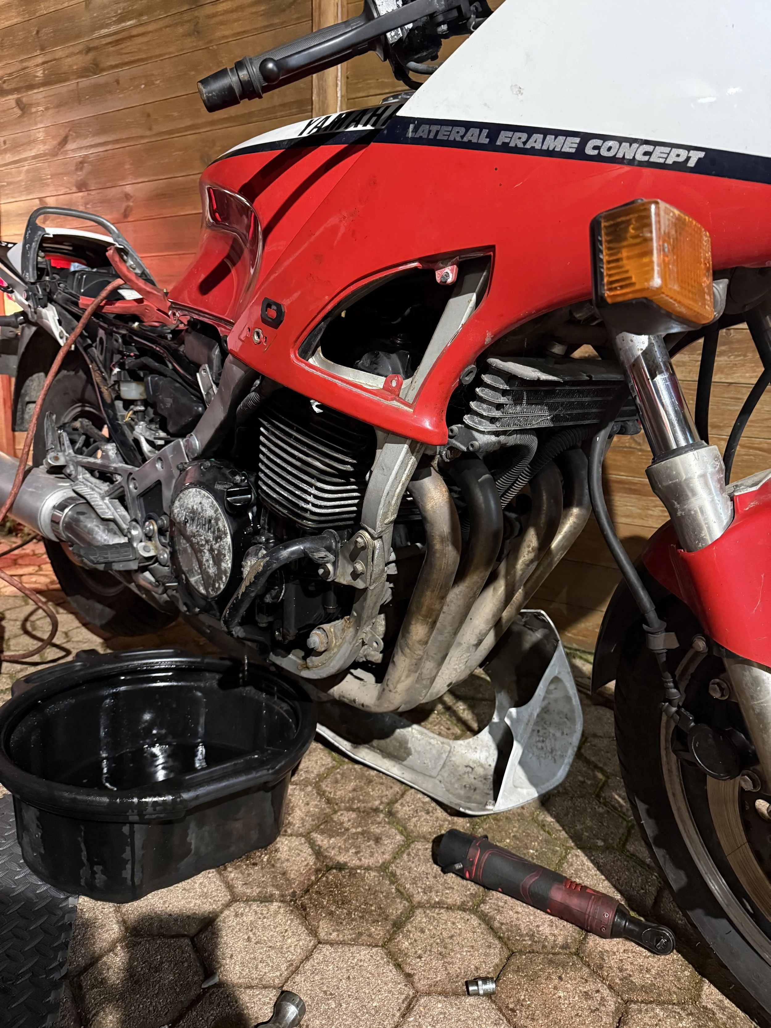 Une moto rouge en cours de réparation, sans carénage avant, avec des outils à proximité, placée sur un pavé. L'intérieur du cadre et le moteur sont visibles.
