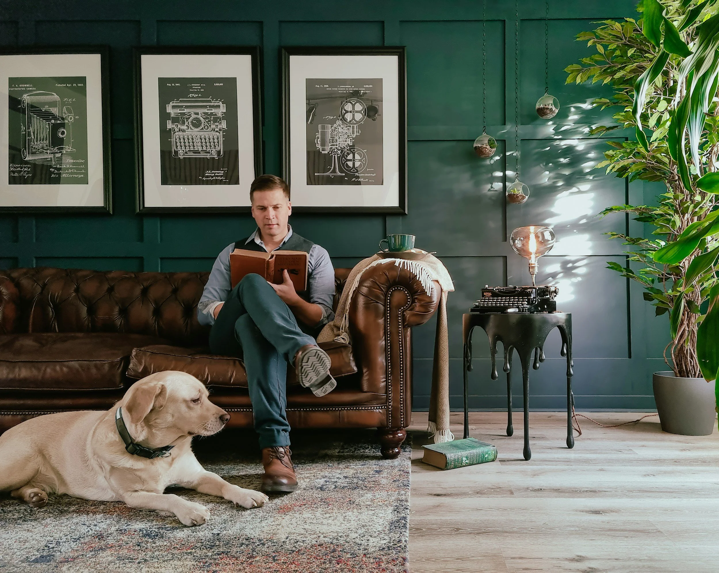 A man sitting on a brown leather couch reading a book, with a yellow Labrador Retriever lying on the rug nearby. The room has dark green walls adorned with framed drawings, plants, and vintage decor.
