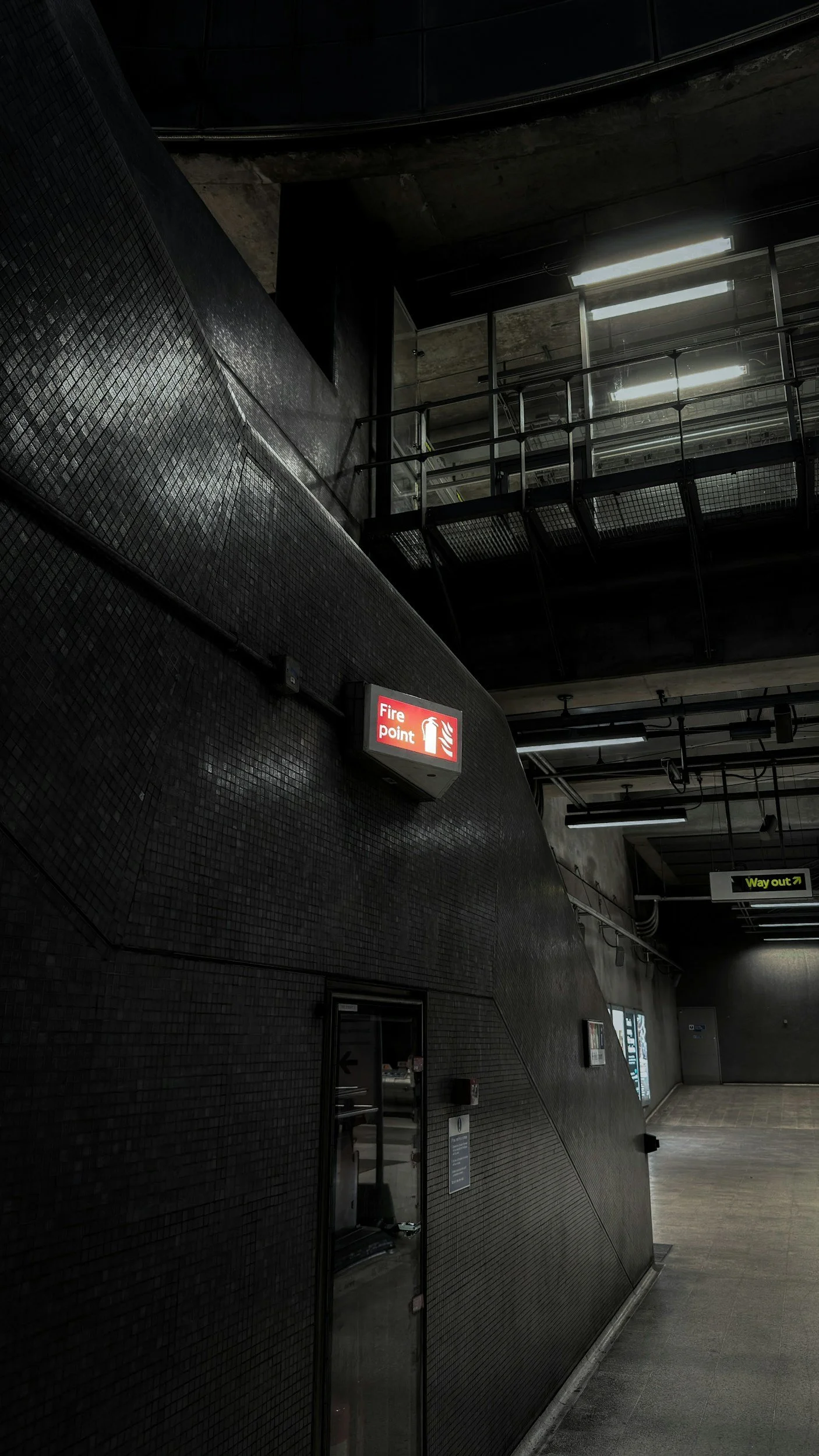 Dark interior of a building with a lit fire point sign on a textured black wall, a glass door, and a hallway leading to an exit sign, metal railing, and industrial-style ceiling lights.