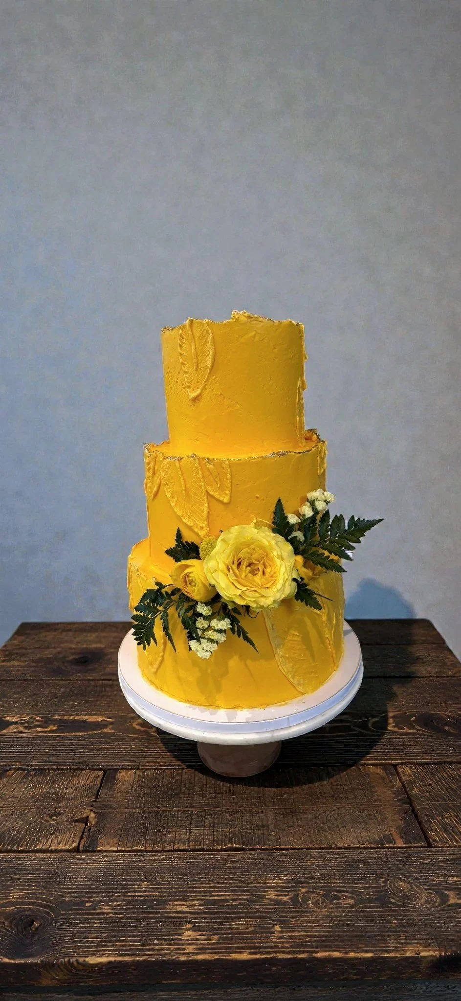 Three-tier yellow wedding cake decorated with yellow flowers and green foliage, placed on a white cake stand on a wooden table.