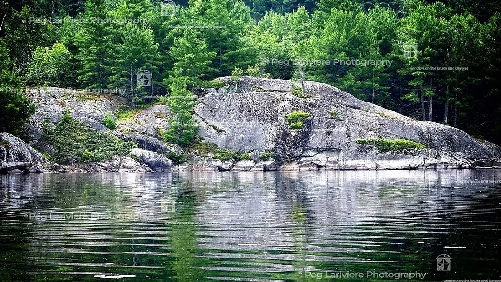 Shoreline of the Serpent Harbour on Lake Huron. The granite grey boulders and brilliant green trees are reflected in the dark water.