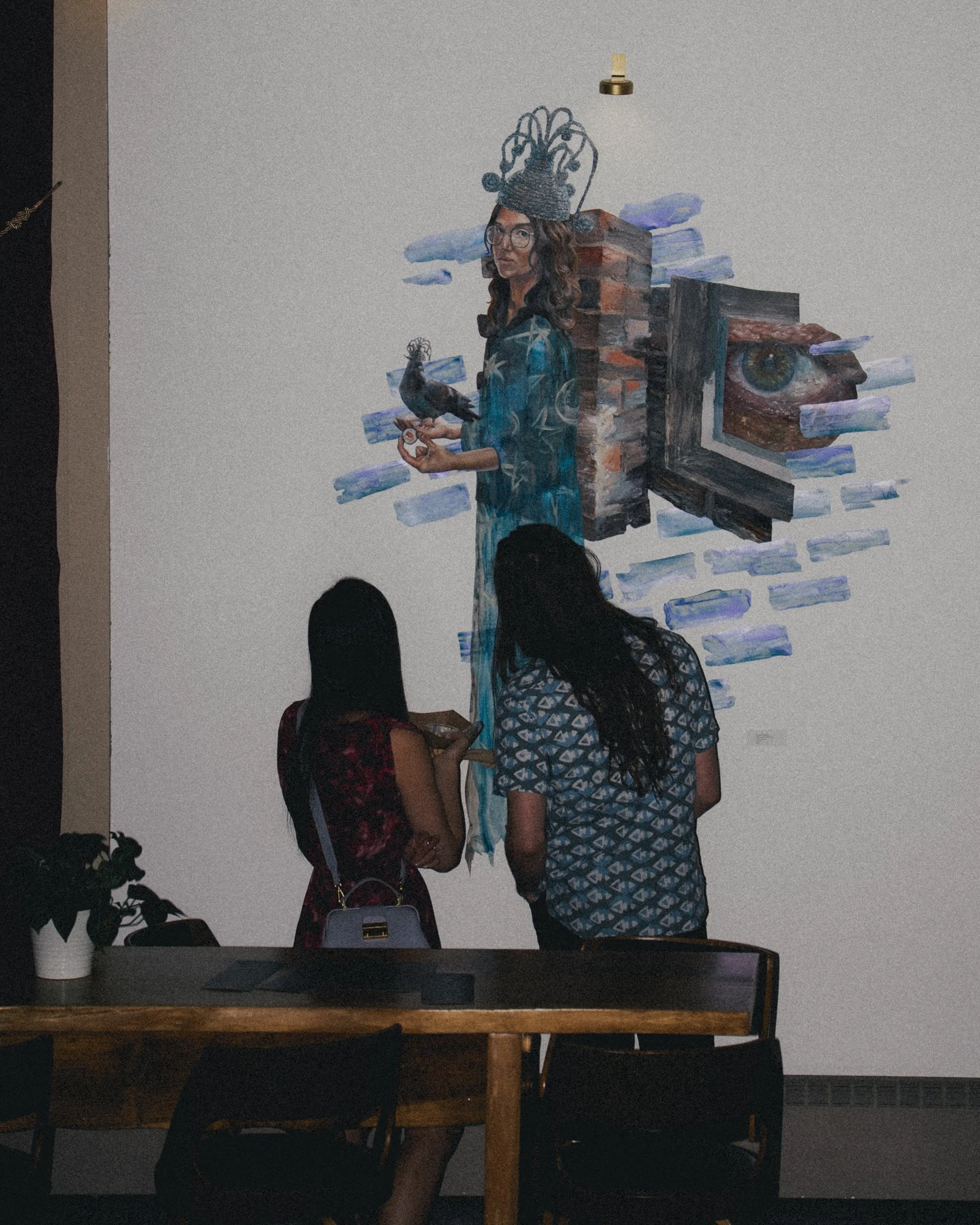 A mural on a white wall depicts a woman with glasses holding a bird, with imaginative elements like a face in a window, colorful clouds, and a key. Two women are observing the artwork in a room with a wooden table and a potted plant.
