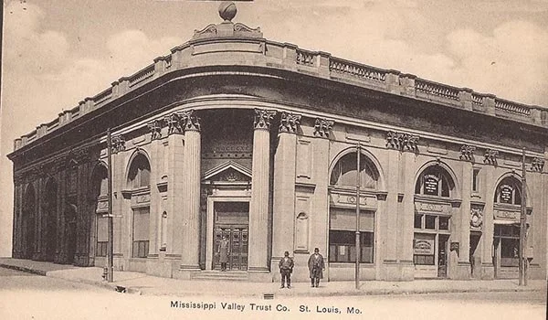 A historic black and white photo of the Mississippi Valley Trust building in St. Louis, Missouri, featuring classical architecture with tall columns and ornate details, with two people walking in front.