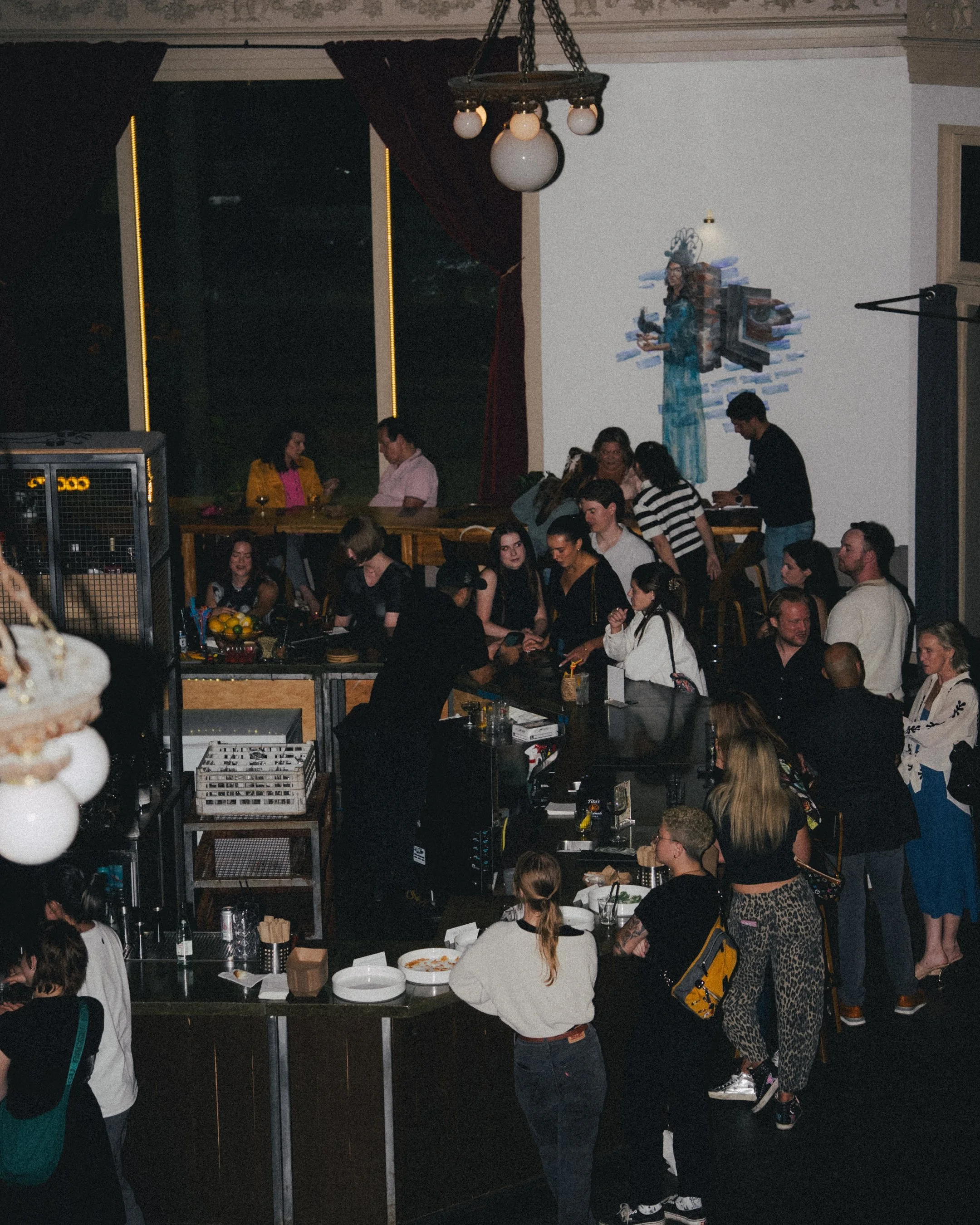 Crowd of people gathered inside a dimly lit bar or restaurant during nighttime, with some standing in line and others sitting at the bar, featuring wall art and hanging light fixtures.