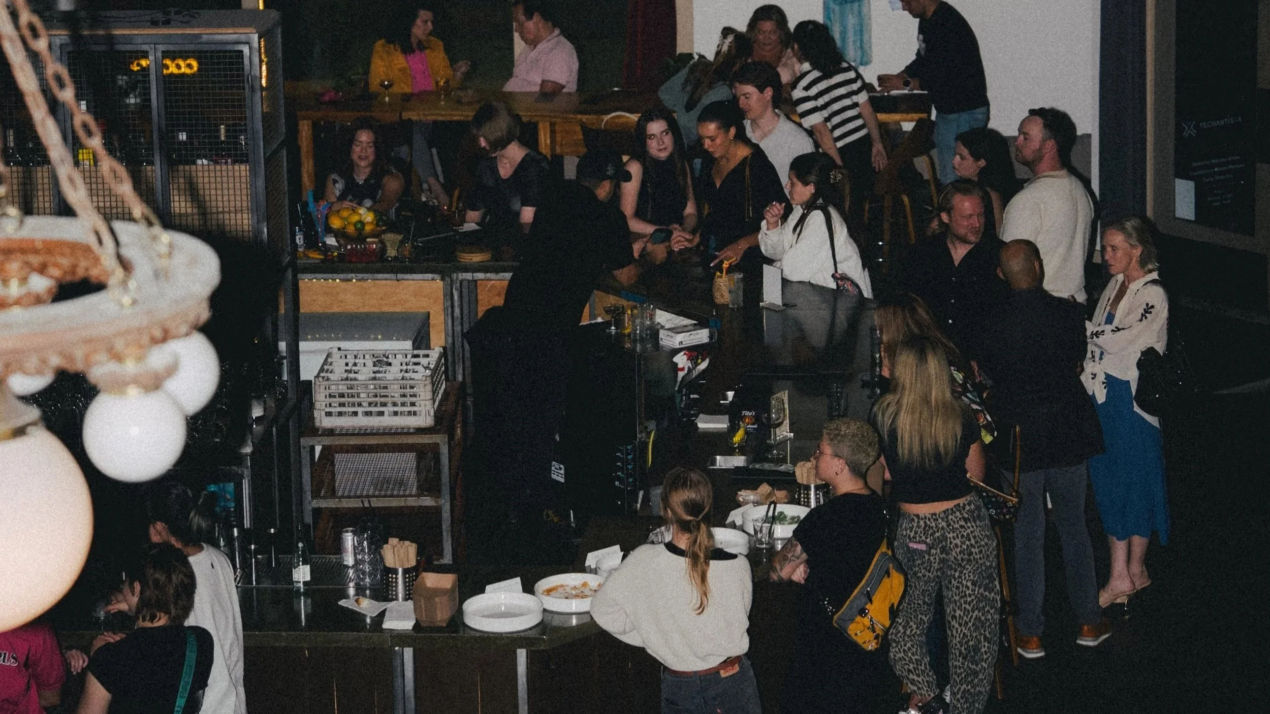 People gathered in a bar or restaurant, some ordering drinks, others socializing and waiting in line. The scene shows a busy indoor setting with various groups of people standing and sitting, some engaged in conversation.