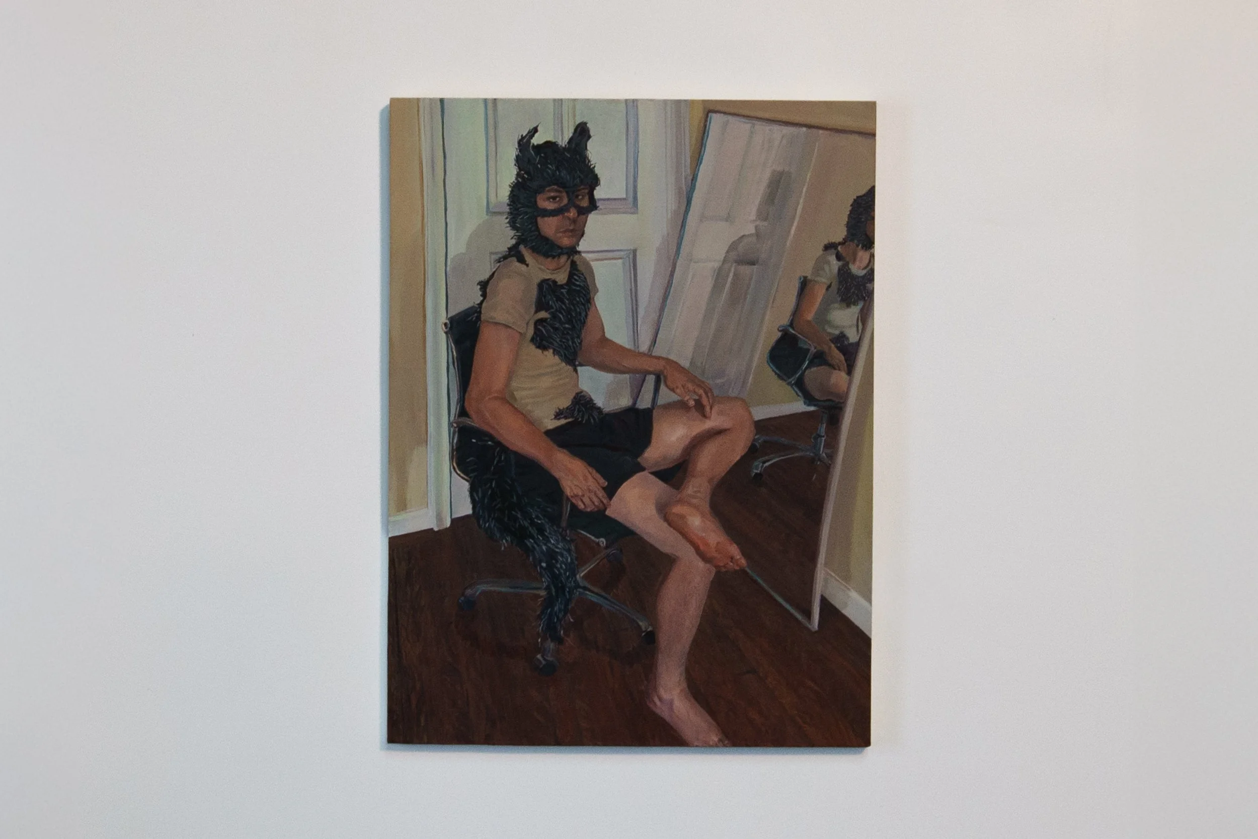 A person wearing a black dog costume with a mask and ears, sitting on a stool, with their legs crossed, in front of a mirror in a room with light-colored walls and dark wooden floor.
