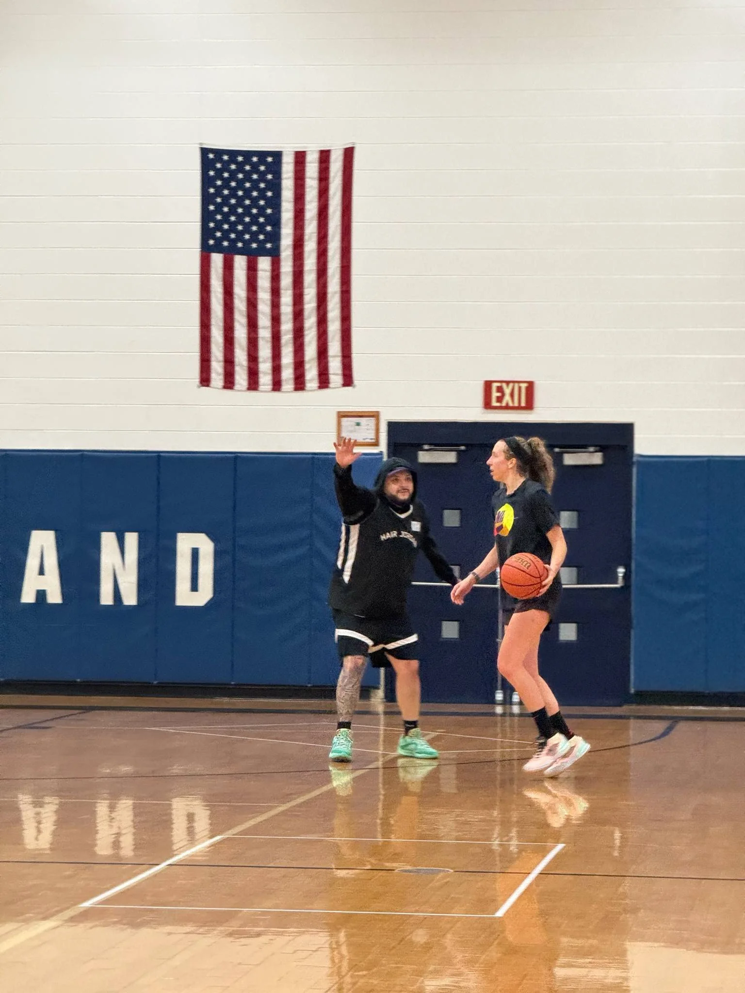 A woman holding a basketball and a man wearing sports attire practicing basketball in an indoor gymnasium with an American flag on the wall.