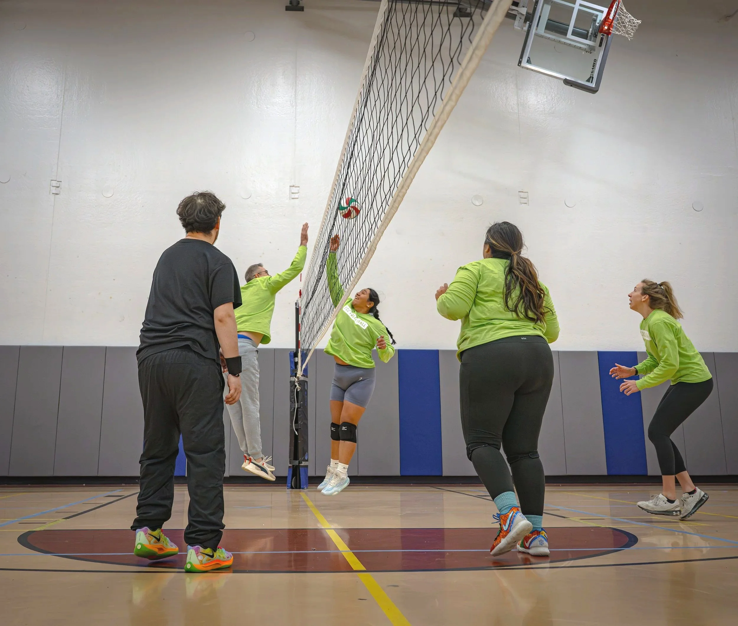 A group of people playing volleyball indoors, with two players jumping near the net to hit the ball while other teammates watch.