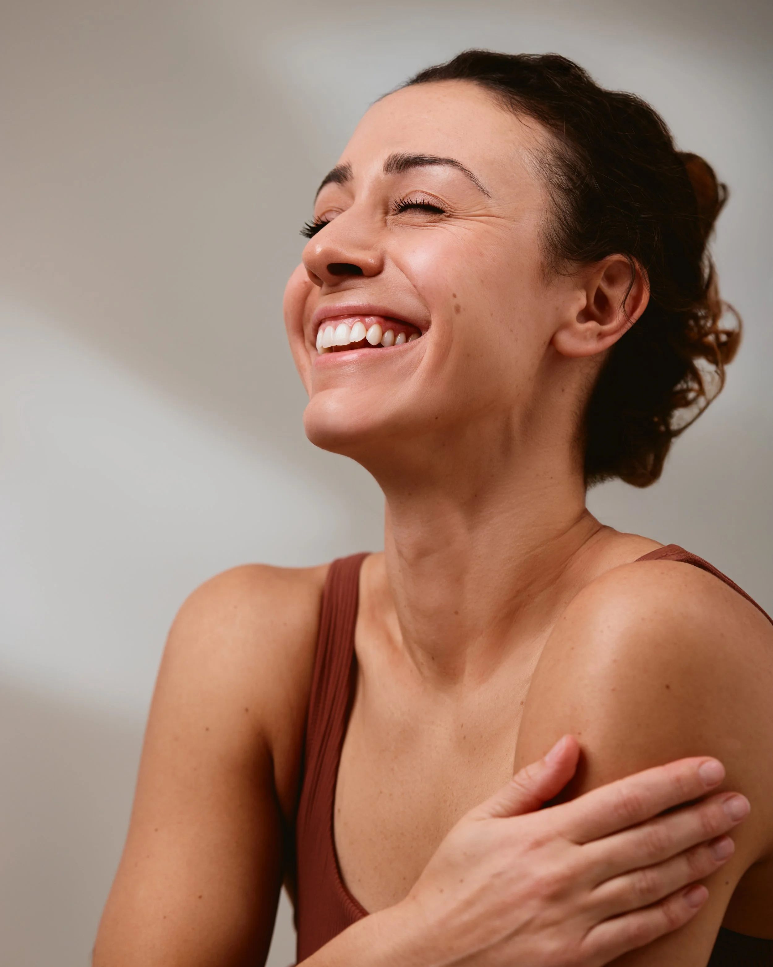 A woman with brown hair smiling with eyes closed, holding her shoulder.