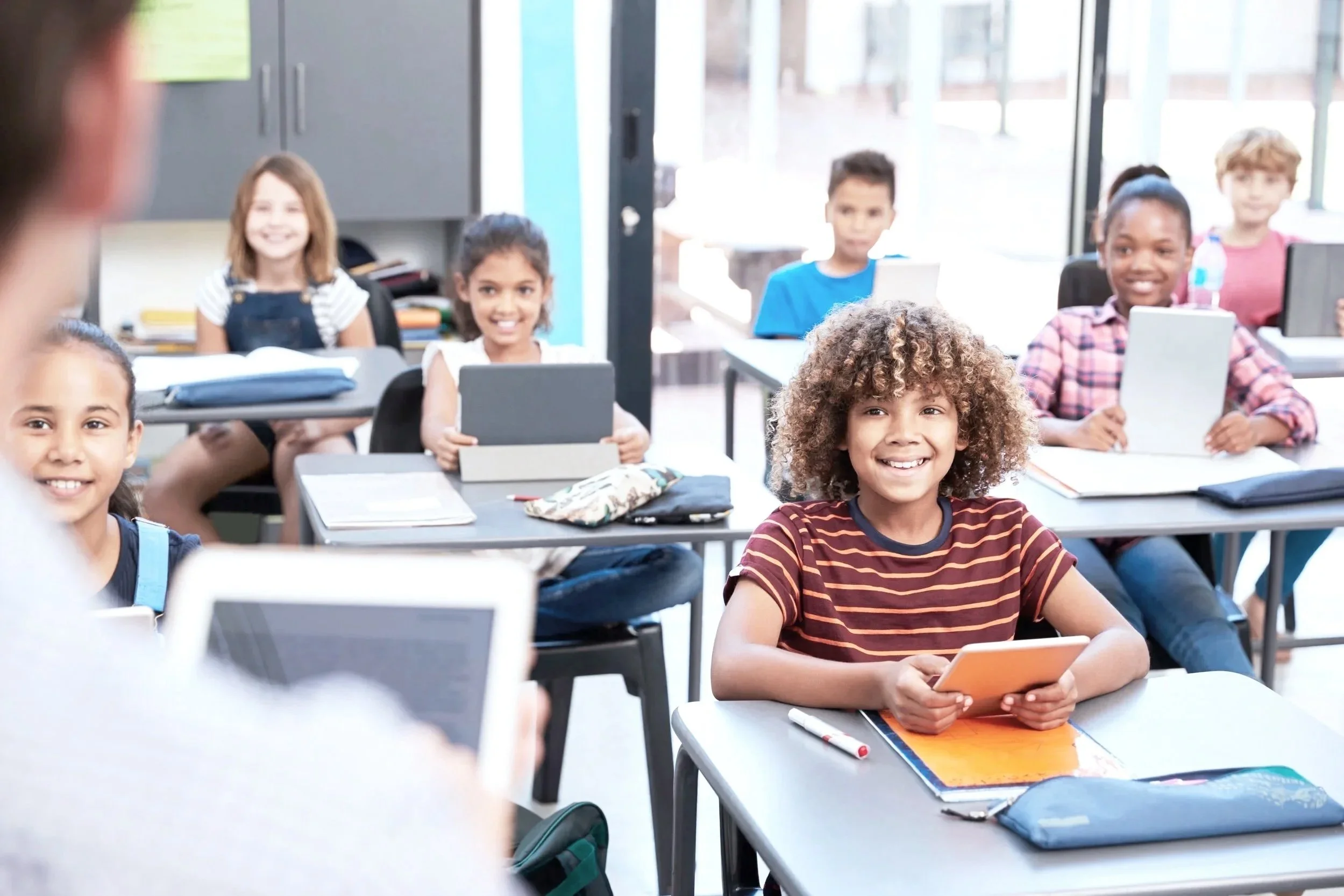 Young students in classroom, smiling and holding tablets, with a teacher in the foreground using a device.