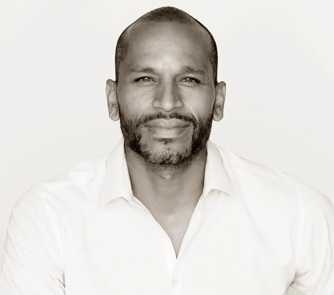 Black and white portrait of a man with short hair and a beard, wearing a white shirt, looking at the camera with a slight smile.