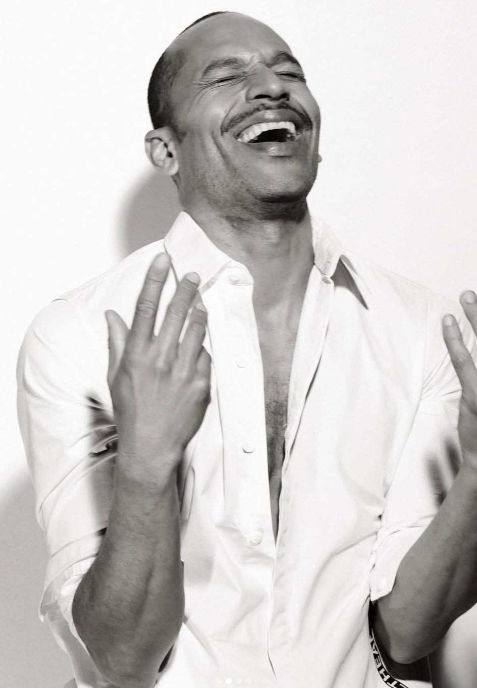 A man with a mustache laughing with his eyes closed, wearing a white shirt with the top buttons undone, against a plain background.