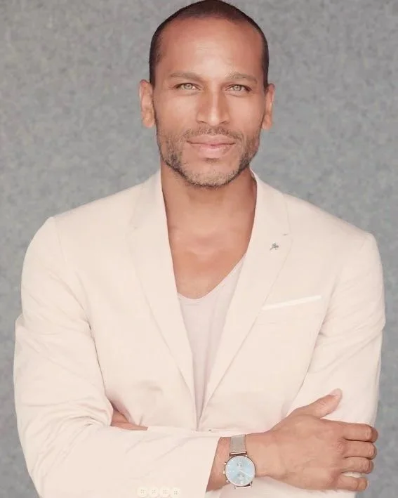A man with short dark hair, light skin, and a beard, wearing a white blazer and light-colored shirt, standing against a gray background.
