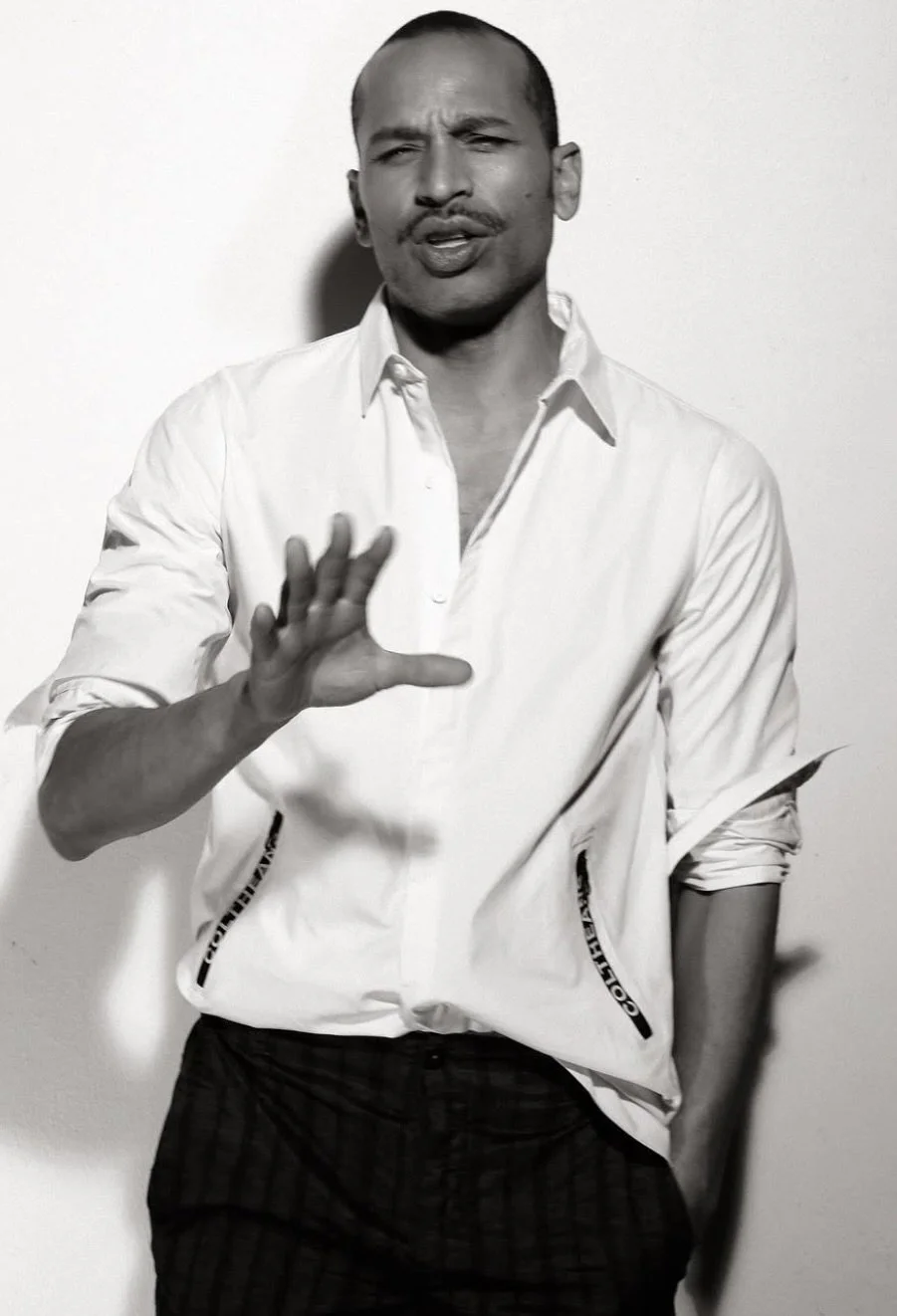 A man with a mustache and a shaved head wearing a white shirt, gesturing with his hand, standing against a plain wall.