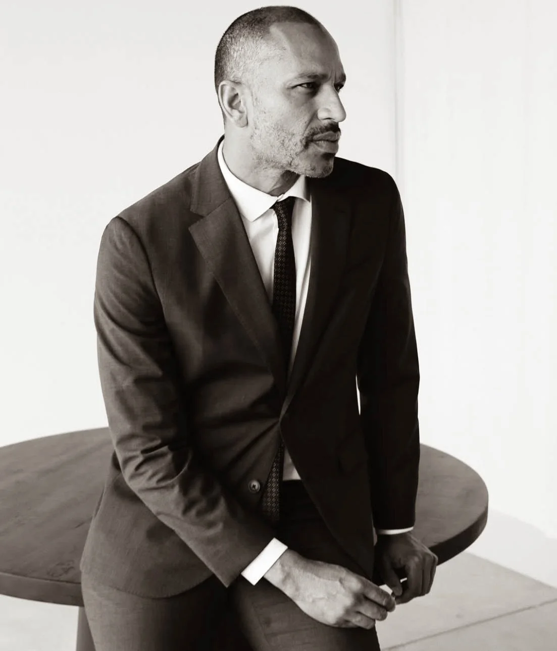 Black and white photo of a man in a suit leaning on a round table against a plain white wall, looking to the right.