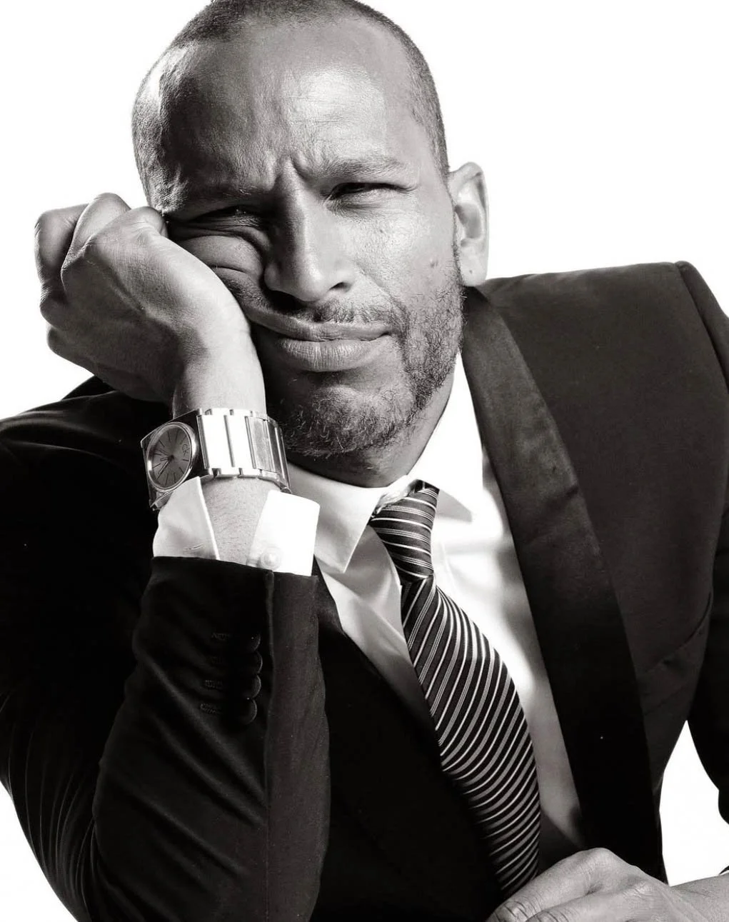 A man in a suit and striped tie, wearing a wristwatch, with his head resting on his hand, making a pained facial expression.