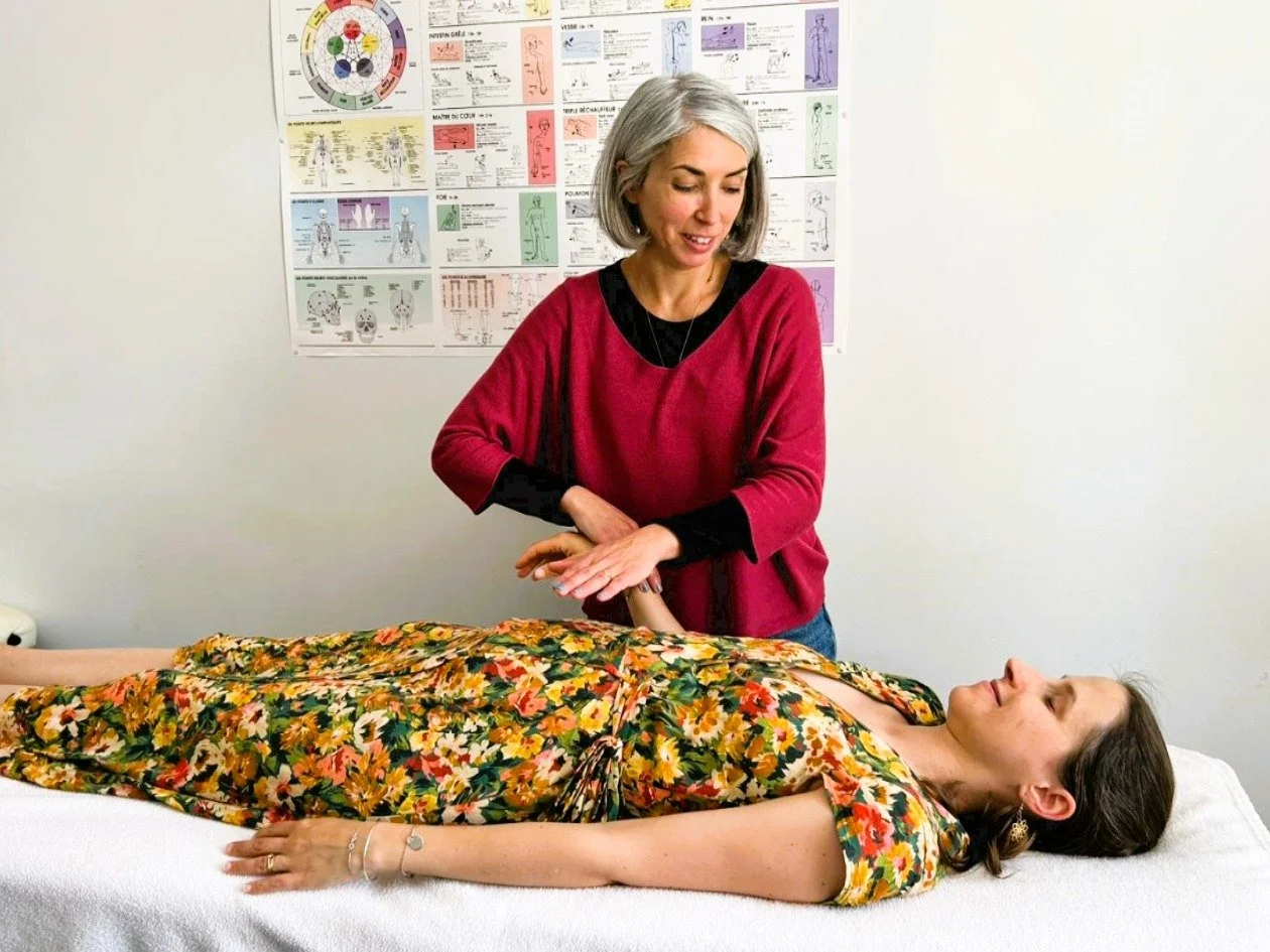 Une femme donne une séance de thérapie ou de soins énergétiques à une autre femme allongée sur une table d'examen, dans une salle avec des posters médicaux en arrière-plan.
