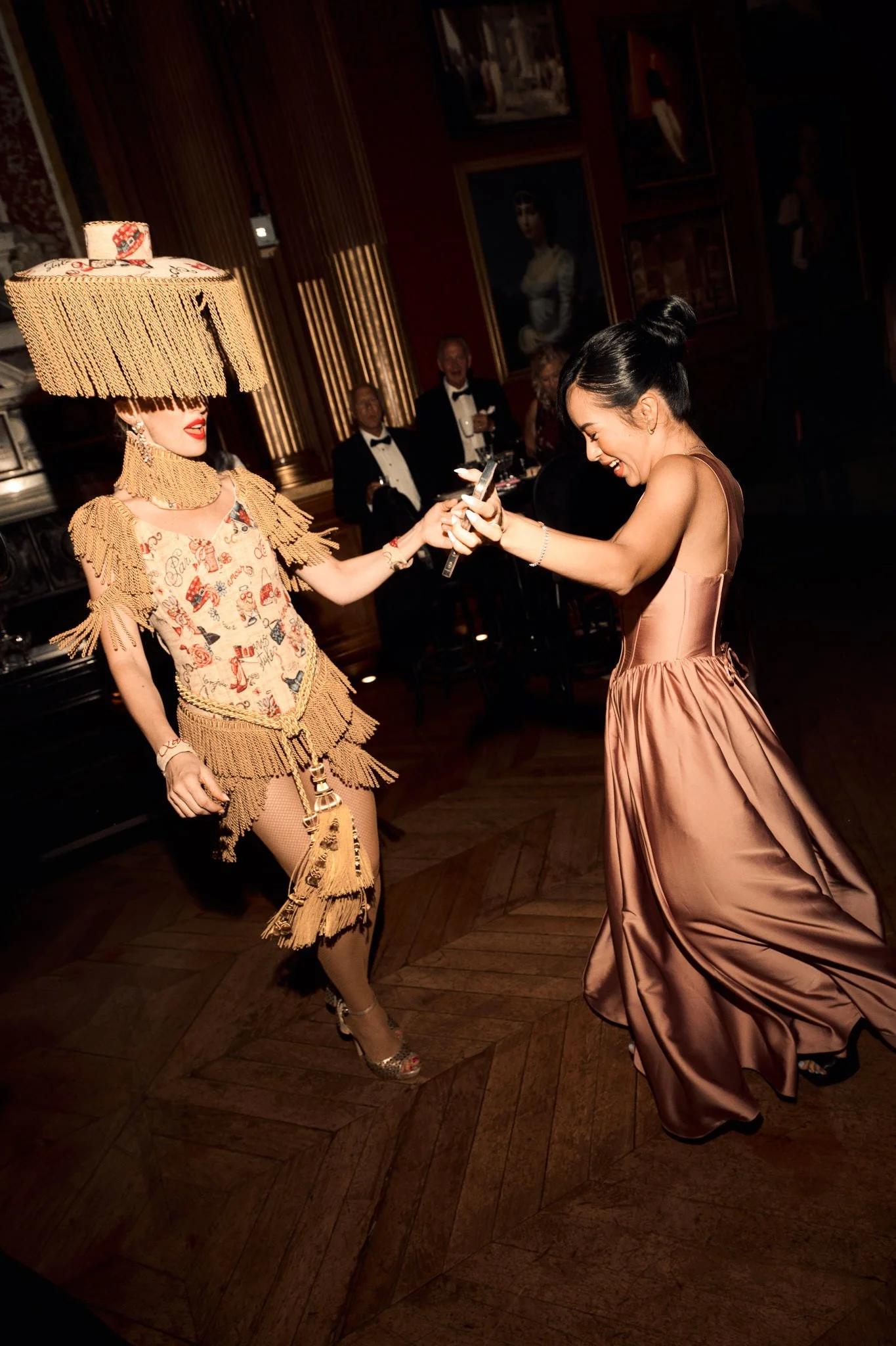 Deux femmes s'amusent lors d'une célébration élégante. L'une porte une robe rose satinée et danse, l'autre porte un costume écossais avec un grand abat-jour en guise de chapeau. Des personnes en smoking sont en arrière-plan dans une salle ornée de po