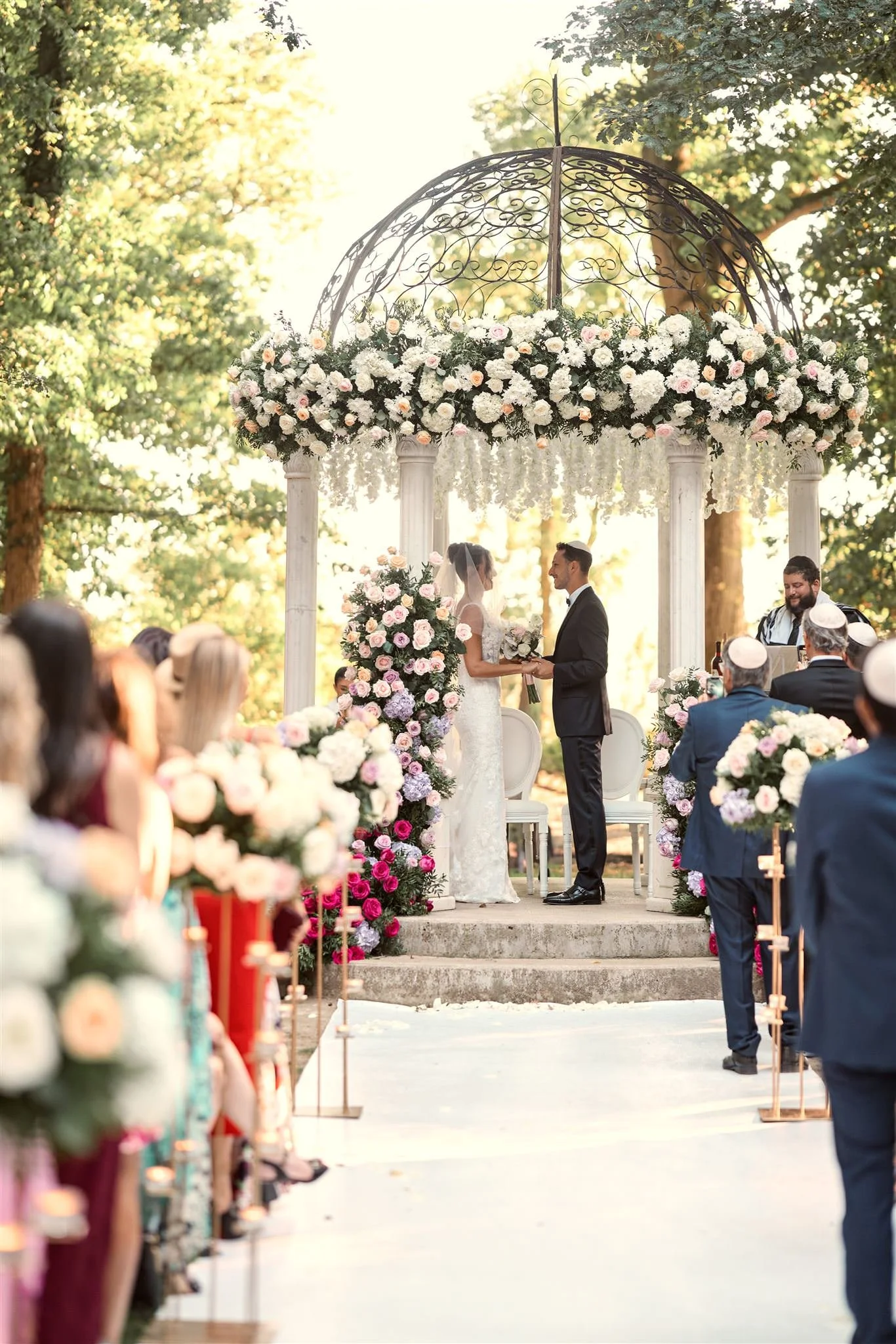 Un couple de mariés échange des vœux lors d'une cérémonie de mariage en plein air sous une structure en fer ornée de fleurs blanches et roses. Audience assise et décorations florales autour. L'événement se déroule dans un environnement boisé en soiré